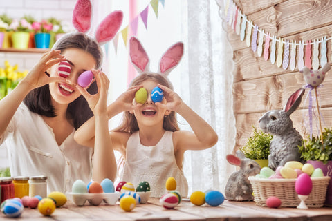 Mother and daughter wearing bunny ears, holding colorful Easter eggs in front of their eyes, surrounded by Easter decorations and a rabbit figurine.