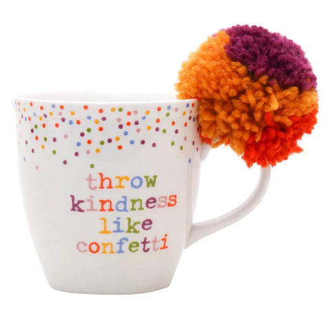 A white mug with colorful polka dots and the phrase "throw kindness like confetti," adorned with a large multicolored pompom.